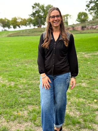 A woman wearing a black long-sleeve button-up blouse with patterned shoulder detailing and blue jeans, standing outdoors in a grassy park.
