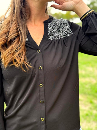 A woman wearing a black long-sleeve button-up blouse with a black and white geometric patterned yoke and gold-tone buttons, photographed outdoors against a blurred green background.