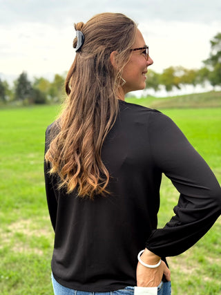 A woman with long brown hair is shown from the back in an outdoor grassy field, wearing a black long-sleeve top and blue jeans.