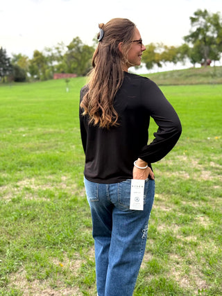 A woman standing outdoors in a grassy field, shown from the back and side. She is wearing a black long-sleeve top and blue jeans, with a white paper tag attached to the back of her jeans.