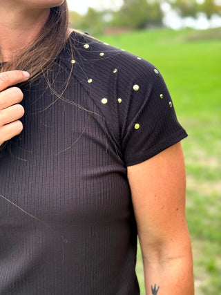 A woman wearing a black short-sleeve t-shirt with small silver rhinestone embellishments on the shoulders and blue jeans, standing in a grassy field.