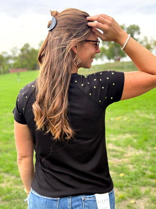 A woman wearing a black short-sleeve t-shirt with small silver rhinestone embellishments on the shoulders and blue jeans, standing in a grassy field.