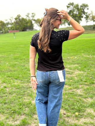A woman wearing a black short-sleeve t-shirt with small silver rhinestone embellishments on the shoulders and blue jeans, standing in a grassy field.