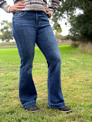 A person wearing dark blue flared denim jeans and a patterned knit sweater, standing outdoors on a grassy field with trees in the background.
