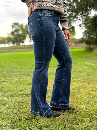A person wearing dark blue flared denim jeans and a patterned knit sweater, standing outdoors on a grassy field with trees in the background.
