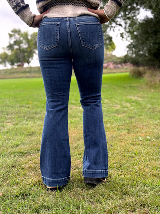 A person wearing dark blue flared denim jeans and a patterned knit sweater, standing outdoors on a grassy field with trees in the background.
