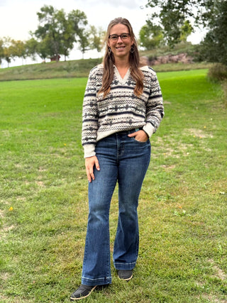 A woman wearing a cream and black patterned knit sweater, blue flared jeans, and black shoes stands on a grassy lawn outdoors.