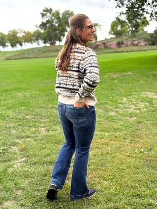 A woman wearing a cream and black patterned knit sweater and blue jeans stands on a grassy field, photographed from the back and side. The background shows a park with trees and a hill under an overcast sky.