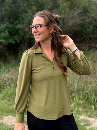 A woman wearing a muted olive green long-sleeve button-up blouse with a collar and chest pocket, standing outdoors in a grassy, wooded setting.