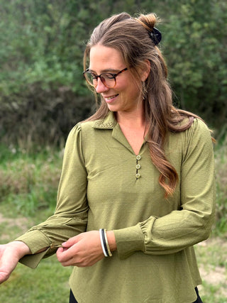 A woman wearing a muted olive green long-sleeve button-up blouse with a collar and chest pocket, standing outdoors in a grassy, wooded setting.