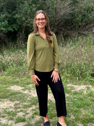 A woman wearing a muted olive green long-sleeve button-up blouse with a collar and chest pocket, standing outdoors in a grassy, wooded setting.