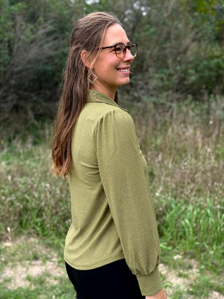 A woman wearing a muted olive green long-sleeve button-up blouse with a collar and chest pocket, standing outdoors in a grassy, wooded setting.
