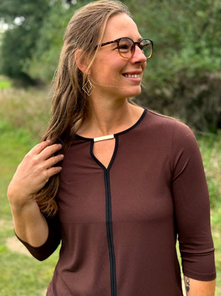 A lady wearing a 3/4 sleeve chocolate top with a gold bar detail at the neckline. 