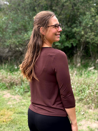 A lady wearing a 3/4 sleeve chocolate top with a gold bar detail at the neckline. 
