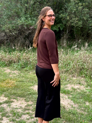 A lady wearing a 3/4 sleeve chocolate top with a gold bar detail at the neckline. 