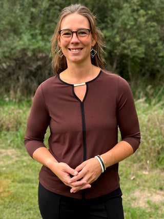 A lady wearing a 3/4 sleeve chocolate top with a gold bar detail at the neckline. 