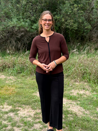 A lady wearing a 3/4 sleeve chocolate top with a gold bar detail at the neckline. 