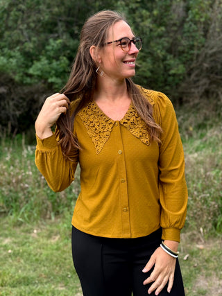 A woman wearing a mustard yellow long-sleeve button-up top with a large textured crochet collar and black pants, standing outdoors in a grassy area with trees in the background.