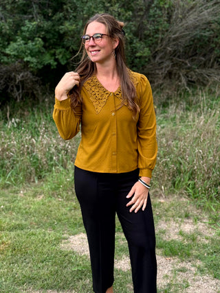 A woman wearing a mustard yellow button-up blouse with a decorative crochet-style yoke and black pants, standing outdoors in a grassy area with trees in the background.