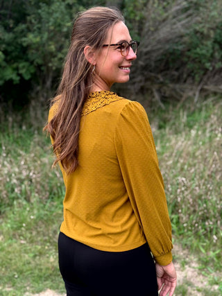 A woman with glasses and long light brown hair is shown from the back and side, wearing a mustard yellow long-sleeve top with a lace yoke and black pants, standing outdoors in a grassy area.