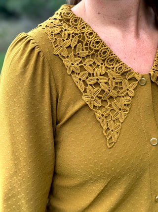 Close-up of a person wearing a mustard yellow blouse with a large floral lace V-neck collar and front buttons.