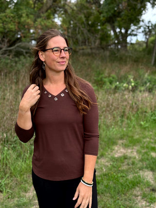 A woman with glasses and long brown hair stands outdoors in a grassy field, wearing a ribbed three-quarter sleeve top in a deep brownish-rose color with decorative silver-tone studs at the neckline, paired with black pants.