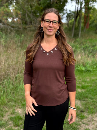 A woman wearing a brown ribbed long-sleeve V-neck top with silver geometric stud embellishments at the neckline, paired with black pants, standing outdoors in a grassy field.