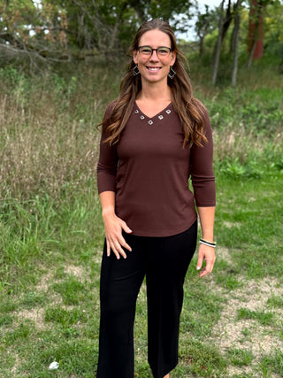 A woman wearing a brown long-sleeve V-neck top with small silver geometric embellishments and black pants, standing outdoors in a grassy field.