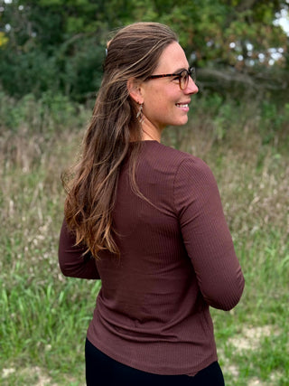 A woman with long brown hair and glasses, wearing a ribbed long-sleeve top in a dark brownish-rose color, standing outdoors in a grassy field.