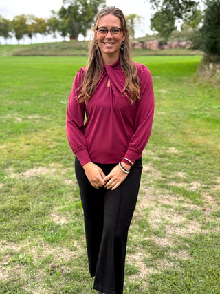 A woman wearing a long-sleeve burgundy mock neck blouse and black wide-leg pants, standing outdoors on a grassy field.