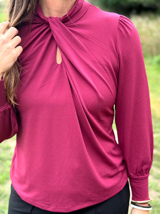 A woman wearing a long-sleeve high-neck blouse in a deep magenta pink, featuring a twisted knot detail at the collar and puff sleeves, photographed outdoors against a blurred green background.