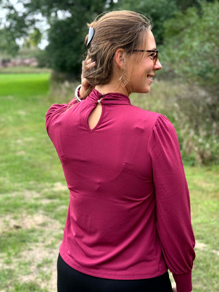 A woman wearing a long-sleeve burgundy mock neck blouse and black wide-leg pants, standing outdoors on a grassy field.