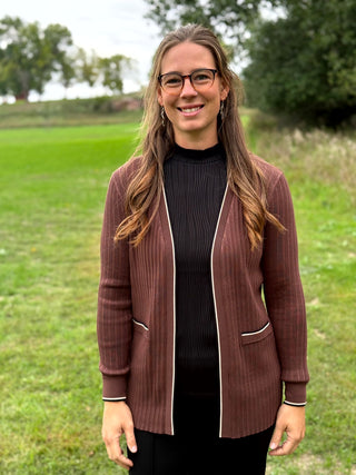 A woman standing outdoors on a grassy field wearing a brown ribbed  cardigan with white piping over a black ribbed top and black pants.