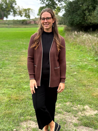 A woman standing outdoors on a grassy field wearing a brown ribbed  cardigan with white piping over a black ribbed top and black pants.