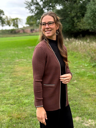 A woman standing outdoors on a grassy field wearing a brown ribbed  cardigan with white piping over a black ribbed top and black pants.