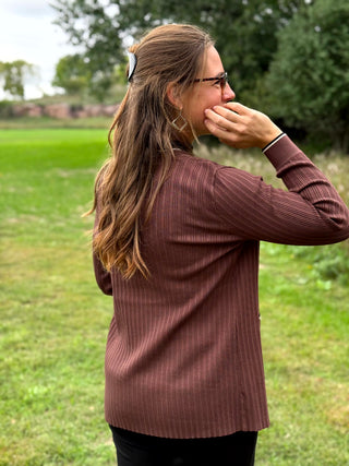 A woman with long brown hair and glasses, wearing a ribbed brown long-sleeve top, standing outdoors in a grassy field.