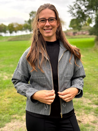 A woman wearing a gray textured zip-front jacket with a drawstring hem over a black top, standing outdoors on a grassy field.