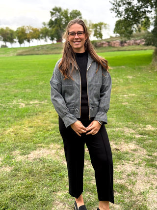 A woman wearing a gray textured zip-up jacket over a black top and black pants, standing outdoors on a grassy field with trees in the background.