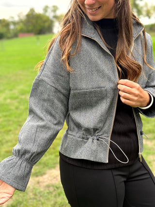 A woman wearing a gray textured zip-front jacket with a drawstring hem over a black top, standing outdoors on a grassy field.