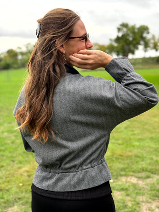 A woman wearing a gray textured long-sleeve jacket and black pants, photographed from behind in an outdoor grassy field.