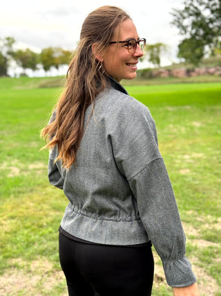 A woman wearing a gray textured long-sleeve jacket and black pants, photographed from behind in an outdoor grassy field.