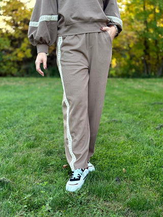 Front walking view of taupe pants with side pockets and cream lace stripes, paired with black-and-white sneakers.