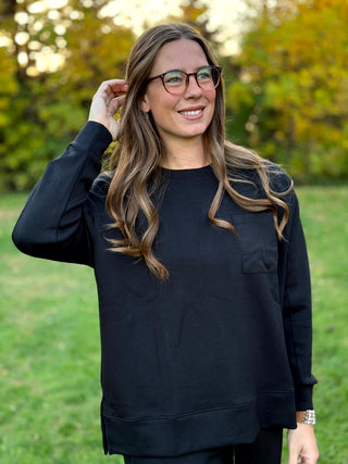 Model wearing a black relaxed-fit crewneck top with long sleeves and a chest pocket, paired with matching wide-leg pants.

