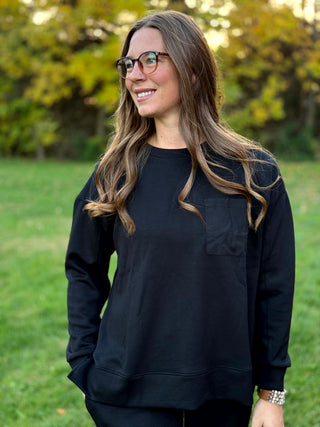  Front view of a model wearing a black crewneck top with a chest pocket and wide hem.