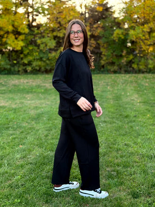 Side view of a model wearing a loose black top and matching wide-leg pants with black-and-white sneakers.