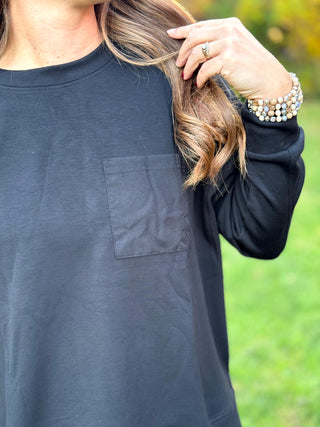 Close-up of a black crewneck top showing the chest patch pocket and long sleeve with cuff.