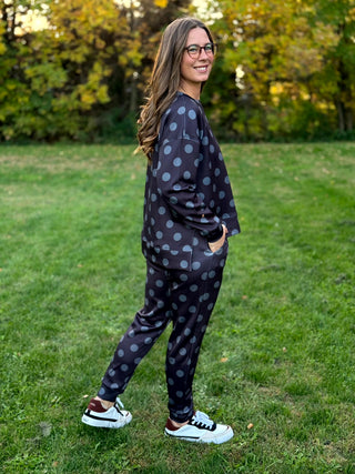 Side view of a model wearing a dark charcoal sweatshirt and matching tapered pants with gray polka dots, hand in side pocket.