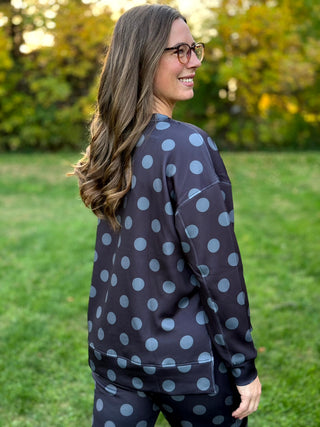 Model wearing a dark charcoal crewneck sweatshirt and matching pants with large gray polka dots, standing outdoors.

