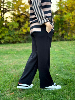 Side view of black wide-leg pants with a hand in the side pocket, styled with white sneakers.