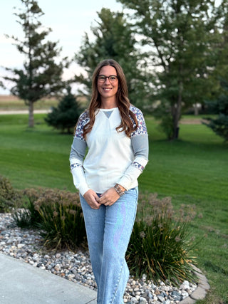 A woman wearing a white long-sleeve top with floral patterned shoulder panels and striped elbow panels, paired with light blue jeans, standing outdoors in a park setting.